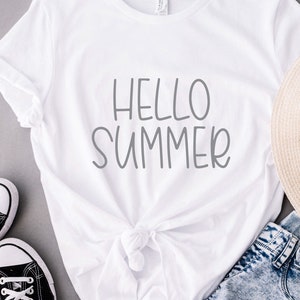 Pode incluir: Camiseta branca com o texto "Hello Summer" em letras cinzas. A camiseta est&aacute; amarrada na cintura e &eacute; combinada com jeans azul, um chap&eacute;u de palha e t&ecirc;nis pretos.