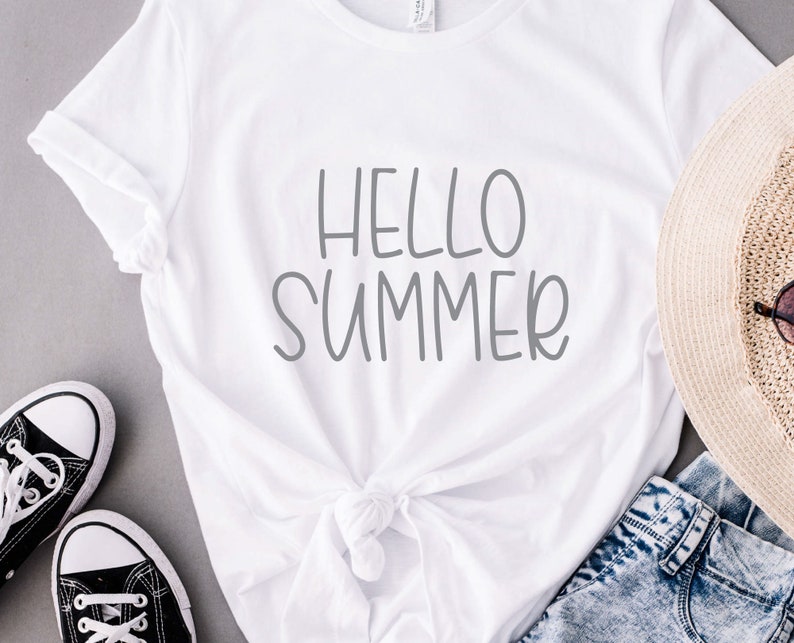 Pode incluir: Camiseta branca com o texto "Hello Summer" em letras cinzas. A camiseta est&aacute; amarrada na cintura e &eacute; combinada com jeans azul, um chap&eacute;u de palha e t&ecirc;nis pretos.