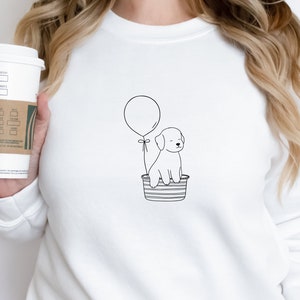 May include: White sweatshirt featuring a black and white line drawing of a puppy in a striped basket, held up by a balloon. The design is simple and minimalist. The sweatshirt is plain and has a round neckline.