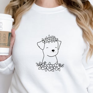 May include: White sweatshirt featuring a black line drawing of a dog wearing a floral crown and surrounded by flowers. The dog has a smiling expression. The sweatshirt is plain and has a crew neck.