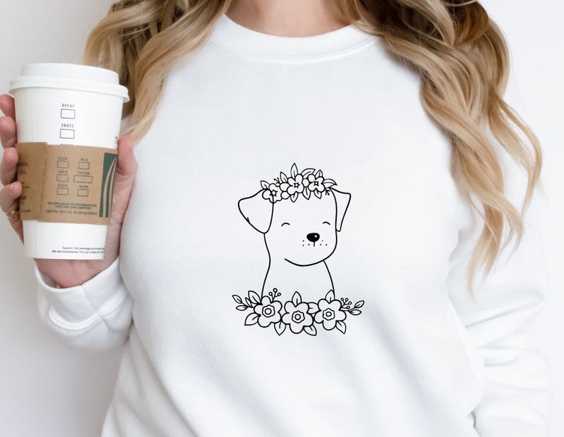 May include: White sweatshirt featuring a black line drawing of a dog wearing a floral crown and surrounded by flowers. The dog has a smiling expression. The sweatshirt is plain and has a crew neck.