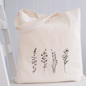 Puede incluir: Una bolsa de lona blanca con un dibujo de l&iacute;nea negra de cinco flores silvestres diferentes. Las flores son simples y delicadas, con tallos y hojas delgadas.