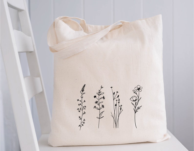 Puede incluir: Una bolsa de lona blanca con un dibujo de l&iacute;nea negra de cinco flores silvestres diferentes. Las flores son simples y delicadas, con tallos y hojas delgadas.