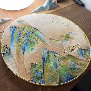 May include: A round, decorative plate with a gold rim features a detailed painting of a mountain range in shades of blue and green. The mountains are depicted in a layered, abstract style, with a flock of birds flying overhead.