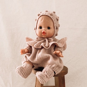 Peut inclure: Une poupée pour bébé vêtue d'un ensemble tricoté rose clair, comprenant un bonnet, une barboteuse et des chaussons. La poupée est assise sur un petit tabouret en bois. La tenue présente un col à volants et des détails en tricot texturé.