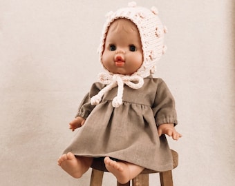 May include: A doll wearing a light pink crocheted hat with a brown dress sitting on a wooden stool.