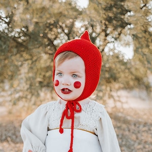 May include: A child in a red knit hood and mushroom-themed costume. The costume includes a white bib with a red mushroom design, a cream-colored blouse, and white leggings. Red Mary Jane shoes complete the outfit.