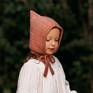 Può includere: Una bambina che indossa un cappello a cappuccio di maglia di colore rosa polvere. Il cappello ha un lungo laccio che è legato sotto il mento. La bambina indossa un vestito bianco e tiene un cesto di fiori.
