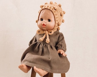 May include: A brown linen dress with a gathered skirt and long sleeves. The dress is worn by a doll with a light brown knitted hat with pom-poms. The doll is sitting on a wooden stool.