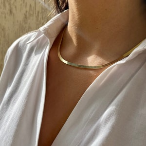 以下が含まれることがあります： シンプルな平らなデザインのゴールドチェーンネックレス。白いシャツを着た人が着用しています。
