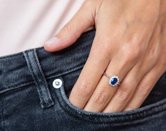 Könnte beinhalten: Ein silberner Ring mit einem ovalen, dunkelblauen Edelstein, umgeben von kleineren weißen Steinen. Der Ring befindet sich an einem Finger, die Hand ruht in einer Jeans-Tasche.