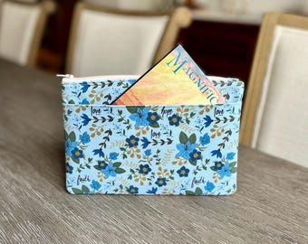 May include: A light blue rectangular zippered pouch featuring a floral pattern in shades of dark blue, green, and gold. The fabric is printed with the words love, hope, and faith. A card with the word MAGNIFIC is tucked into the front pocket.