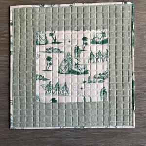 May include: A green quilted placemat with a white center featuring a vintage illustration of the nativity scene. The illustration includes camels, stars, and palm trees.