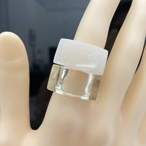 Op de afbeelding: Een witte en transparante ring met een glinsterende afwerking. De ring is gemaakt van twee delen, een wit en een transparant. De ring wordt gedragen aan een hand.