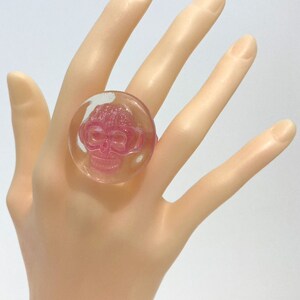 May include: A clear pink ring with a pink skull design. The ring is on a mannequin hand.