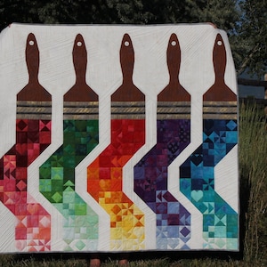 Peut inclure: Une courtepointe colorée représentant six pinceaux faits de carrés de tissu. La courtepointe est blanche avec un manche de pinceau brun et une brosse de pinceau colorée. Les poils du pinceau sont de différentes couleurs, notamment le rouge, l'orange, le jaune, le vert, le bleu et le violet.