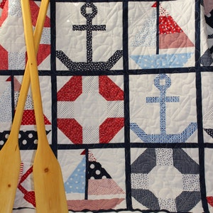 May include: A nautical themed quilt with a patchwork design featuring anchors, sailboats, and life preservers in red, white, and blue. The quilt is displayed on a beige surface with two wooden oars.