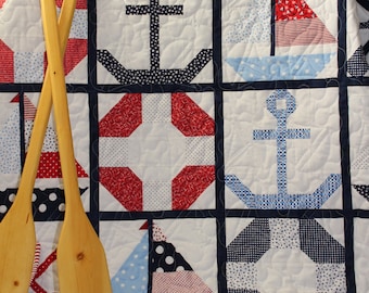 May include: A nautical themed quilt with a patchwork design featuring anchors, sailboats, and life preservers in red, white, and blue. The quilt is displayed on a beige surface with two wooden oars.