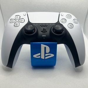 Könnte beinhalten: Ein weißer PlayStation 5 DualSense Wireless-Controller mit einem blauen PlayStation-Logo-Ständer. Der Controller hat schwarze Akzente und Tasten.