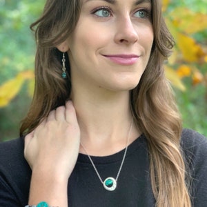 May include: A woman wearing a black shirt with a silver necklace and a green stone pendant. She is also wearing a bracelet with a green stone and a silver charm.