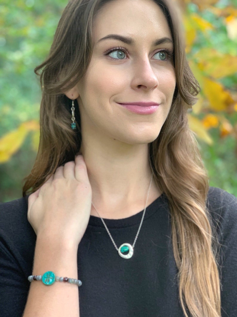 May include: A woman wearing a black shirt with a silver necklace and a green stone pendant. She is also wearing a bracelet with a green stone and a silver charm.