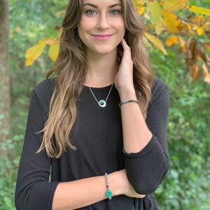May include: A woman wearing a black long-sleeved shirt with a silver necklace and a green stone pendant. She is also wearing a bracelet with a green stone charm.