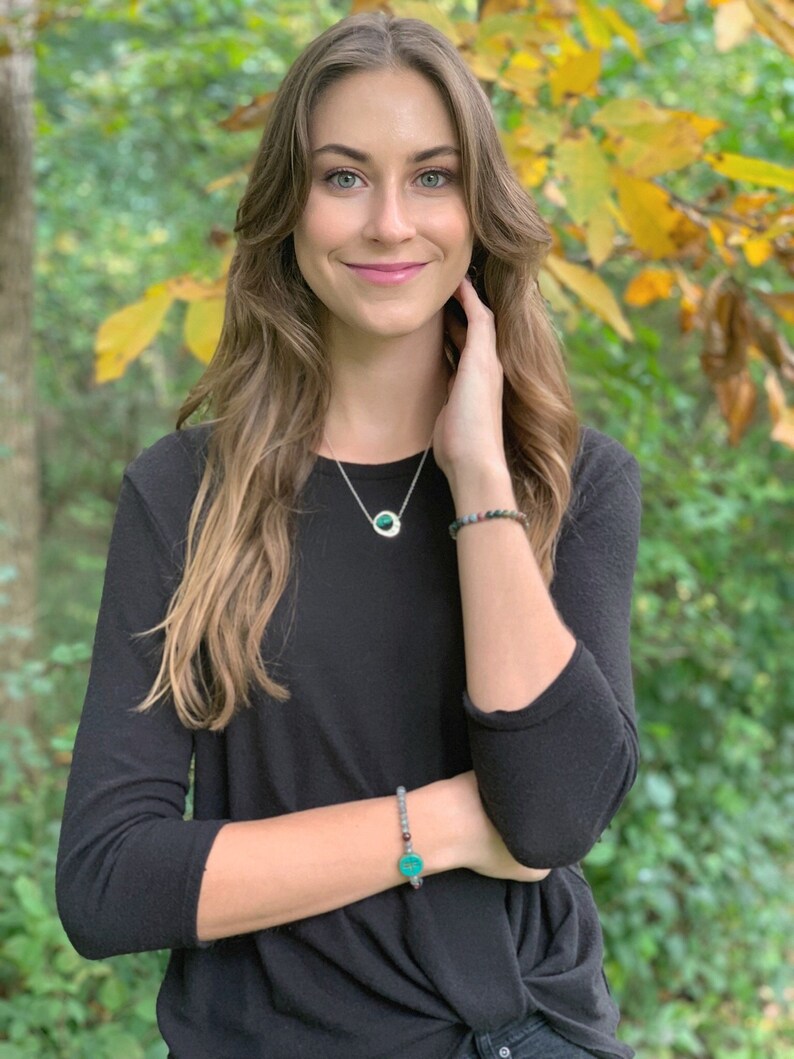 May include: A woman wearing a black long-sleeved shirt with a silver necklace and a green stone pendant. She is also wearing a bracelet with a green stone charm.