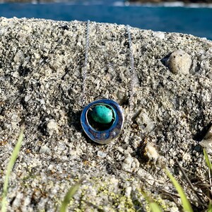 May include: A silver necklace with a turquoise stone set in a circular pendant. The necklace is on a rocky surface with green grass in the foreground.