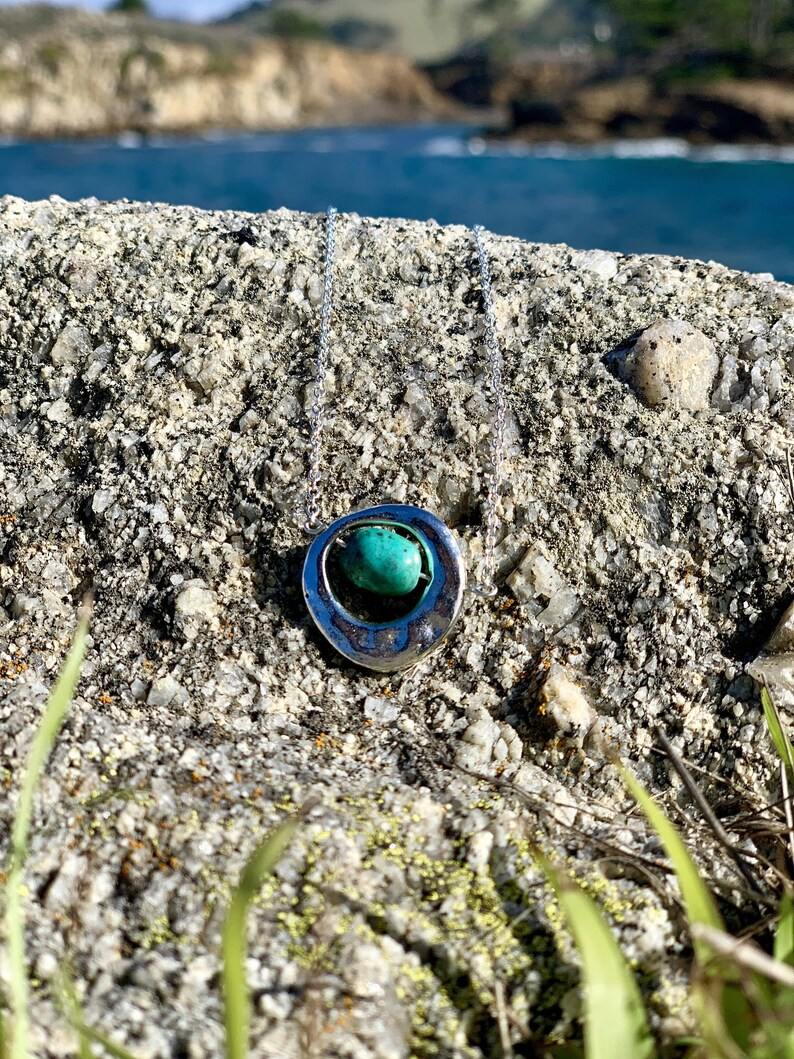 May include: A silver necklace with a turquoise stone set in a circular pendant. The necklace is on a rocky surface with green grass in the foreground.