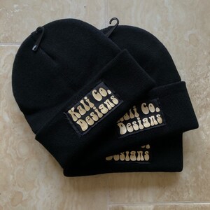 Puede incluir: Dos gorros de punto negros con un logotipo dorado que dice "Kali Co. Designs" en cada gorro.