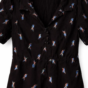 Könnte beinhalten: Schwarzer Overall mit Kragen und kurzen Ärmeln. Der Stoff ist mit bunten Vogelillustrationen versehen. Der Overall hat eine Knopfleiste vorne und eine geraffte Taille, die eine schmeichelhafte Silhouette erzeugt.