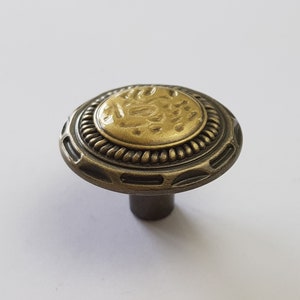 May include: A round, antique-style cabinet knob with a gold-colored center and a dark bronze outer ring. The center has a decorative design.