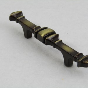 Könnte beinhalten: Antik-Stil Schrankgriff mit Bronze-Finish. Der Griff hat einen rechteckigen Mittelteil und gebogene Stützen. Das Design ist verziert und dekorativ, geeignet für die Möbelrestaurierung oder um einen Vintage-Touch zu verleihen. Ca. 15 cm lang.