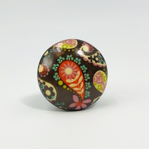 May include: A round cabinet knob with a dark brown base featuring a colorful paisley and floral pattern. The design includes shades of orange, pink, light green, and teal, with small white flower accents distributed across the surface.