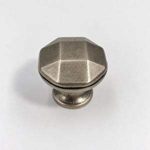 Op de afbeelding: Een meubelknop met een achthoekige bovenkant en een ronde voet, voorzien van een metallic zilveren afwerking met een verweerde, geborstelde textuur. Het beslag heeft een gefacetteerd ontwerp aan de bovenzijde en een gladde, ronde steel.