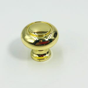 May include: A polished gold-tone metal cabinet knob featuring a smooth, rounded, mushroom-shaped head with a raised circular center detail and a tapered cylindrical base, reflecting light across its shiny, reflective, metallic surface.