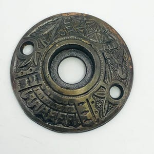 May include: Antique bronze-colored door plate with intricate embossed patterns. The circular plate features a central hole, two smaller holes, and detailed designs around the edges. The plate has a weathered appearance.