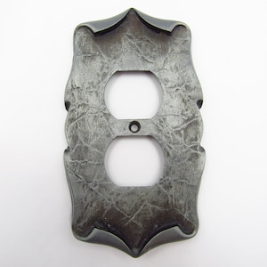 Könnte beinhalten: Eine silberne Steckdosenabdeckung aus Metall mit einem dekorativen, verzierten Design. Die Abdeckung hat eine strukturierte, gehämmerte Oberfläche und zwei Standard-Steckdosenöffnungen.