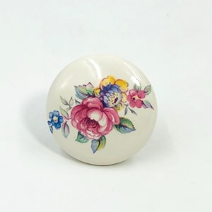 May include: A round, off-white ceramic cabinet knob featuring a colorful floral pattern. The design includes a prominent central pink rose, smaller blue and yellow blossoms, and green leaves painted across the smooth, glossy surface of the knob.