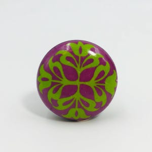 May include: A round cabinet knob featuring a vibrant purple base color with a symmetrical, stylized floral pattern in bright lime green. The design consists of four repeating leaf-like shapes radiating from the center of the circular surface.
