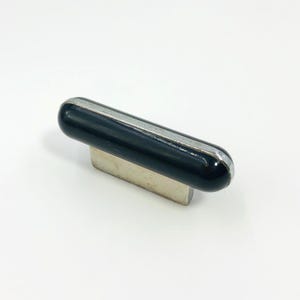 May include: A cabinet knob featuring a cylindrical, glossy black horizontal bar with a metallic silver trim along the top edge. The handle is supported by a rectangular, brushed silver base that extends downward to provide a mounting point.