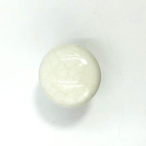 May include: A round, off-white ceramic cabinet knob featuring a glossy finish with a visible network of fine, dark crackle glaze patterns across its surface. The knob has a smooth, domed top and a simple, circular shape for furniture attachment.