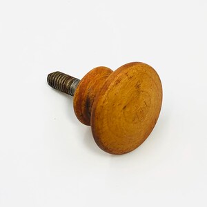 Könnte beinhalten: Ein hölzerner Schrankknopf mit einer runden, flachen Oberfläche und einer Gewindeschraube aus Metall. Der Knopf hat eine warme, hellbraune Farbe, und die Schraube ist ein dunkles, metallisches Grau. Der Knopf ist wahrscheinlich für eine Schublade oder einen Schrank.