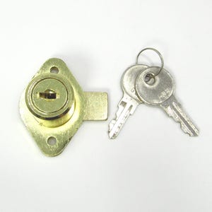 Puede incluir: Una cerradura de armario de color latón con una placa frontal en forma de diamante y dos llaves plateadas en un anillo. La cerradura tiene un ojo de cerradura en el centro y dos orificios para tornillos. Las llaves tienen un diseño simple y funcional.