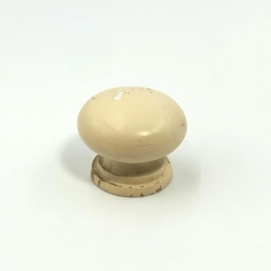 May include: A single, cream-coloured cabinet knob in a mushroom shape. It features a rounded top and a short, cylindrical base with a slightly distressed finish. The knob is likely made of wood or a similar material.