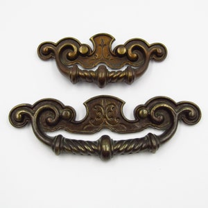 May include: Two antique brass cabinet pulls with ornate scrollwork and a rope-like design. The larger pull is 4 inches wide and the smaller pull is 3 inches wide.