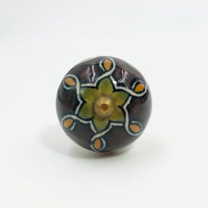 May include: A round ceramic cabinet knob with a dark brown glossy finish. The surface features a central yellow-green star-like floral pattern outlined in white, with small orange petal accents extending outward and a brass-colored metal center.