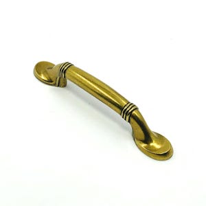 May include: A brass-toned cabinet pull with a curved handle and oval-shaped ends. The handle features decorative bands near the ends. The hardware is designed for furniture and cabinetry.