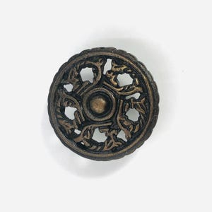 Può includere: Un bottone circolare in metallo color bronzo caratterizzato da un intricato design traforato, una cupola centrale in rilievo e un bordo esterno smerlato. La superficie presenta una finitura scura e vissuta con dettagli in rilievo.