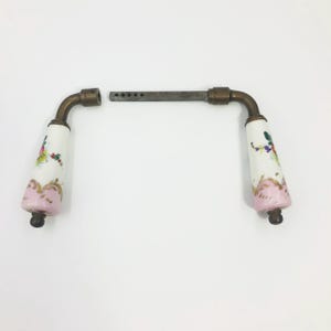 May include: A pair of door handles featuring white porcelain grips with hand-painted floral motifs in pink, green, and yellow, accented by gold-toned trim. The handles are connected by a central metal spindle with a series of small circular holes.